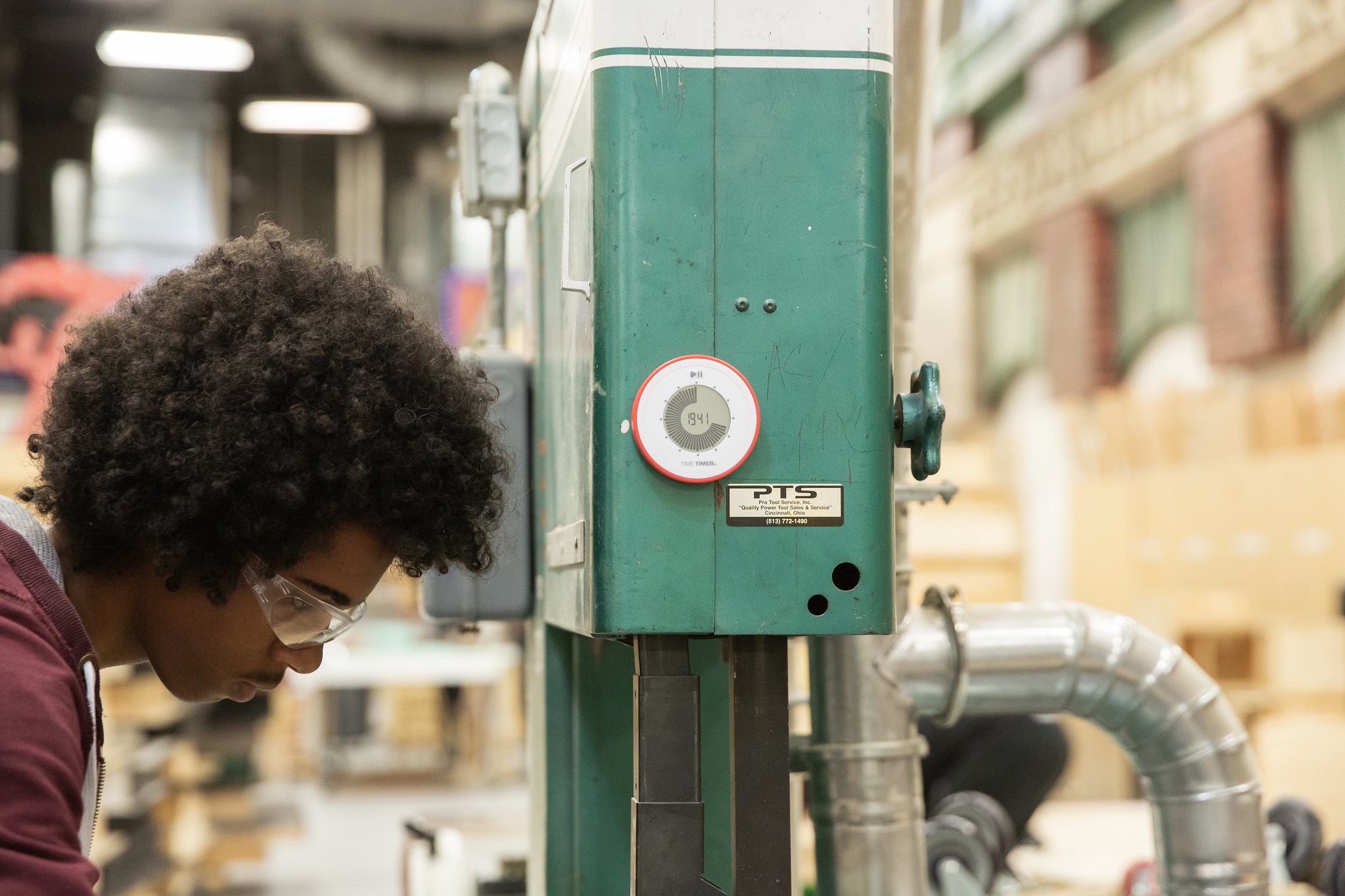 A Time Timer Twist is fixed to a metal box in a shop-like environment. A person with a dark skin tone and a black afro is wearing safety goggles and performing a task out of frame right next to the box.