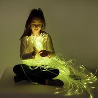 A child sits in a dimly lit room. They have long dark hair and are looking down at a handful of Fiber Optic Tails in their hands. The tails are lit up and make the child's face glow.