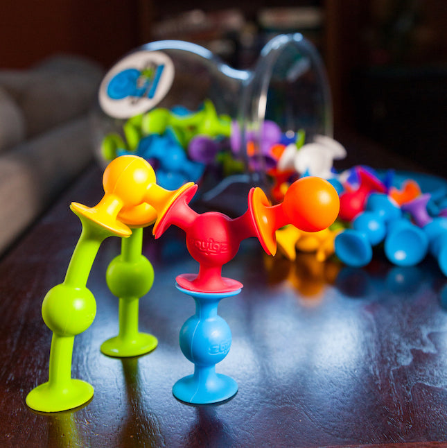 A container for the Deluxe Squigz is on its side in the background and Squigz spill out of it. Several connected Squigz are in the foreground.