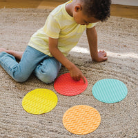 A child with dark skin tone and short brown hair sits over four Sensory Mats. Their forefinger on their right hand is extended and they trace the bumps on the red mat.