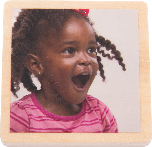 A wood tile with a picture of a dark skinned toddler with long spiraling hair, a pink shirt, and pierced ears is looking off to the side with their mouth open and a small smile.