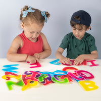 Two children with light skin tone, one with pigtails and one wearing a backwards hat, trace the Textured Gel letters in front of them.