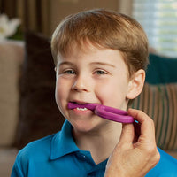 A child with light skin tone and short brown hair is looking into the camera. There is an adult's hand extended off camera, and it is holding up the Magenta Grabber Original to the mouth of the child, who has it between their teeth.