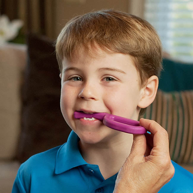 A child with light skin tone and short brown hair is looking into the camera. There is an adult's hand extended off camera, and it is holding up the Magenta Grabber Original to the mouth of the child, who has it between their teeth.