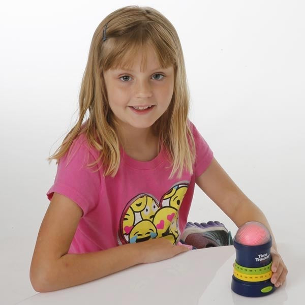 A child with light skin tone and shoulder length blonde hair looks at the camera and smiles. One arm is on the table in front of them and the other is holding onto the base of the Time Tracker Mini.