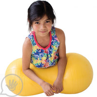A child with medium light skin tone and long brown hair pulled back lies on their belly on a yellow Peanut Ball.