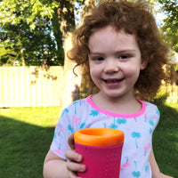 A toddler with light skin tone, wavy brown hair, and brown eyes. They are holding the orange/hot pink cup and smiling while standing in a backyard.