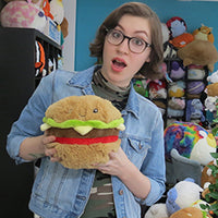 A person with light skin tone and shoulder length brown hair is wearing glasses and has their mouth open in a small smile. They are holding up the Mini Squishable Hamburger.