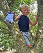 A child with light skin tone and short blonde hair is standing in a tree. They are wearing the black Simply Sleeveless Sensory Compression Shirt with grey shorts. They are smiling and holding a blue stuffed animal.