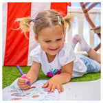 A child with light skin tone and blonde pigtails is holding a pink Pencil Grip on a purple crayon. They are laying on grass and coloring in a picture.