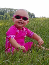 A baby with light skin tone sits in the grass outside wearing the pink petal Bubzee Polarized Wrap Around glasses.