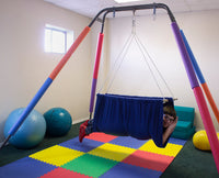 A child with light skin tone and short brown hair lies on their belly in the Soft Taco Jr. Swing Seat. They are in a colorful room surrounded by physical therapy equipment.