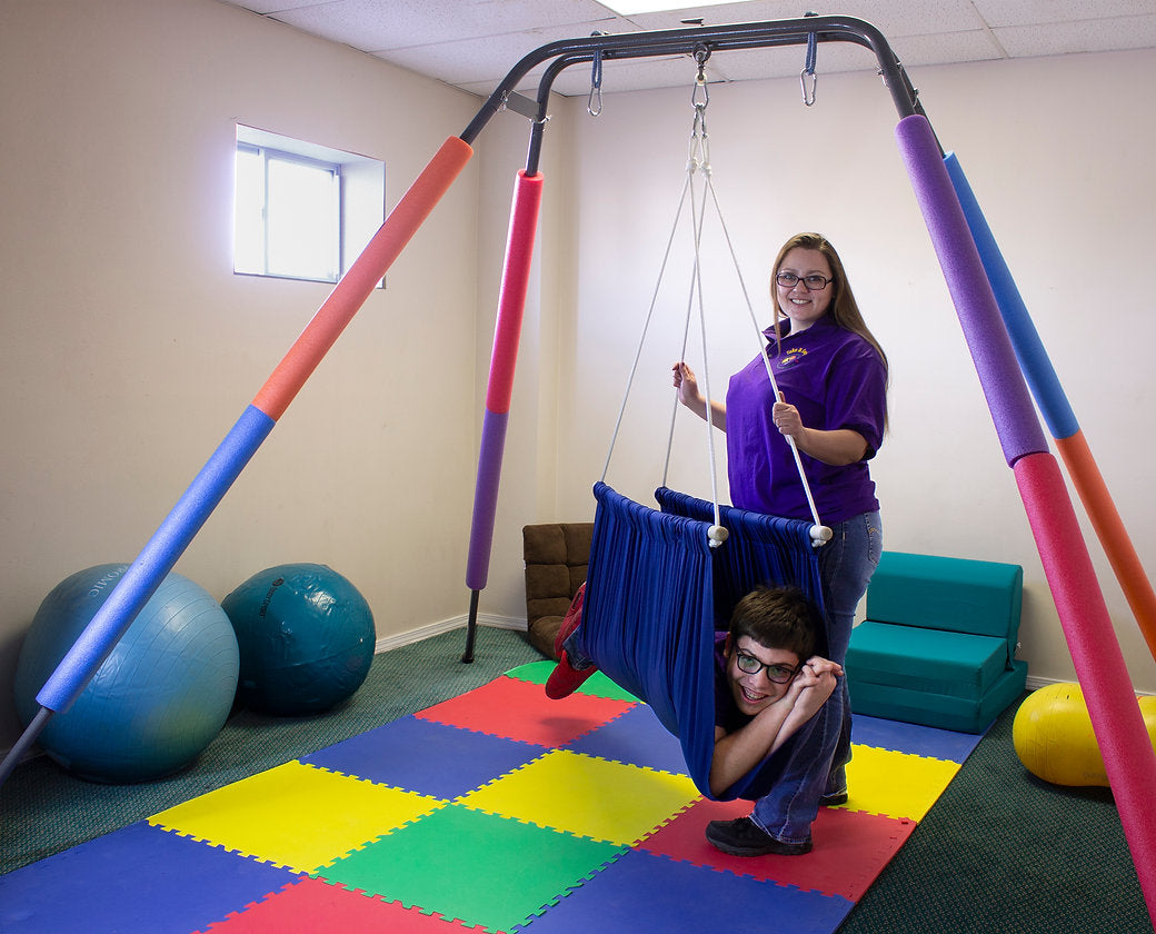 A child with light skin tone and short brown hair lies belly down in the Soft Taco Jr. Swing Seat. A person with light skin tone and long brown hair stands next to them and holds two of the ropes connecting the swing to the stand. They are both in a colorful room surrounded by physical therapy equipment.